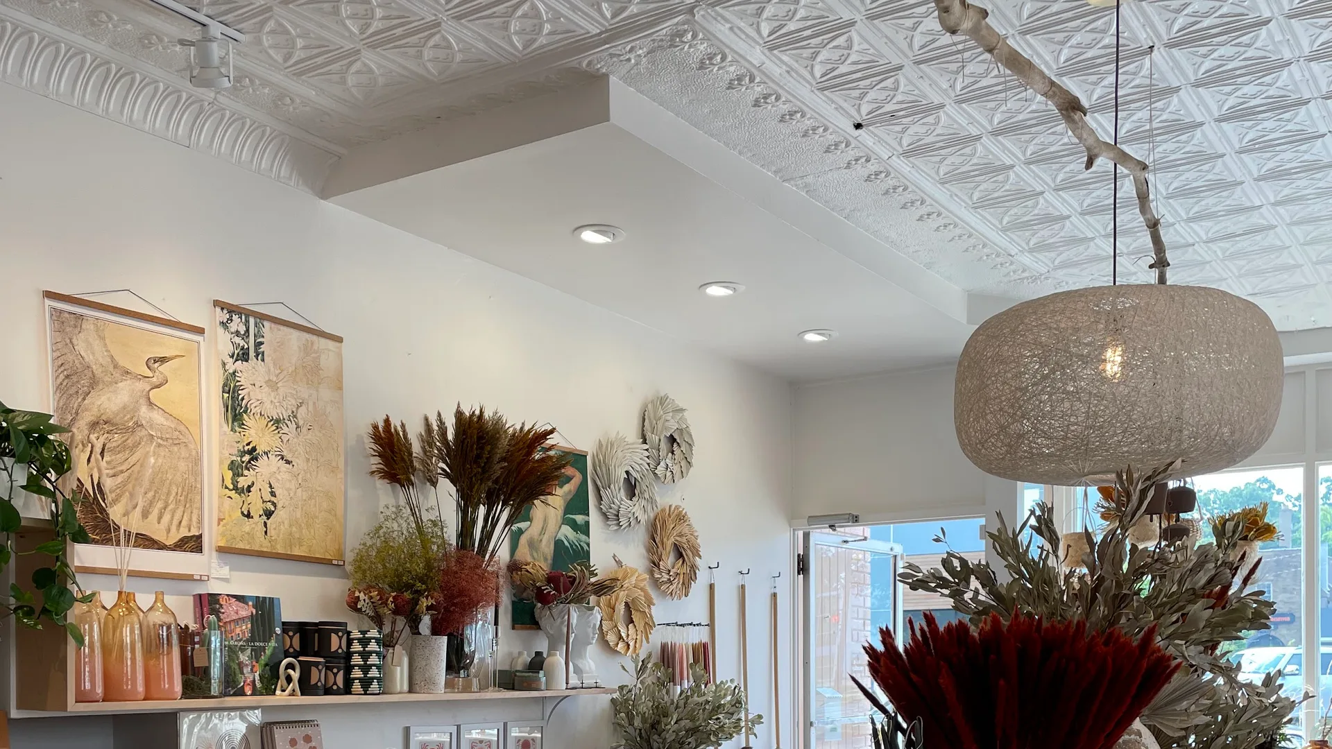 Inside Commune: a whitewashed pressed-tin ceiling above open shelves of candles, ceramics and books, jute rugs over marble floors, and dried florals near the Cherry Street door.
